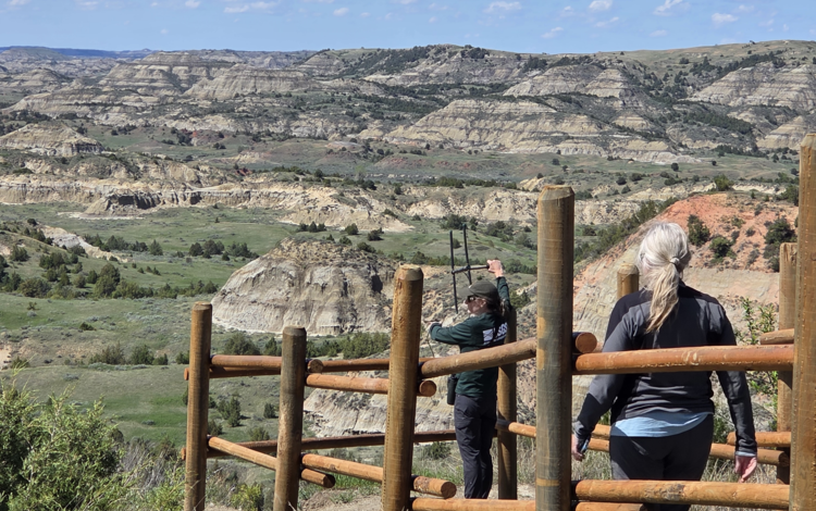 two people stand behind a fence, one holding up a telemetry device, mountains and sky in the background