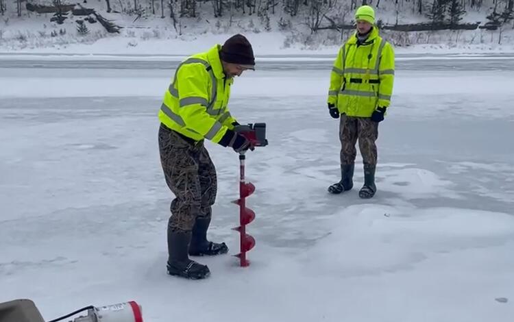 Two scientists standing on a frozen river drilling a hole to check the flow below
