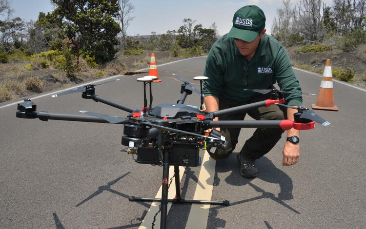 a man in dark green USGS gear kneels next to a large black drone in the middle of a road, cones and trees in the background