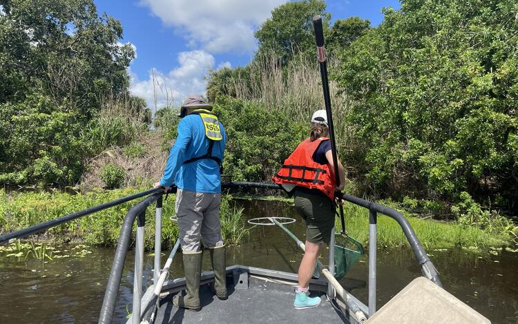Two people in life jackets stand on a bow platform of a boat while electrofishing.