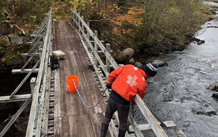 Hydrologic Technician leaning over bridge railing collecting water quality sample