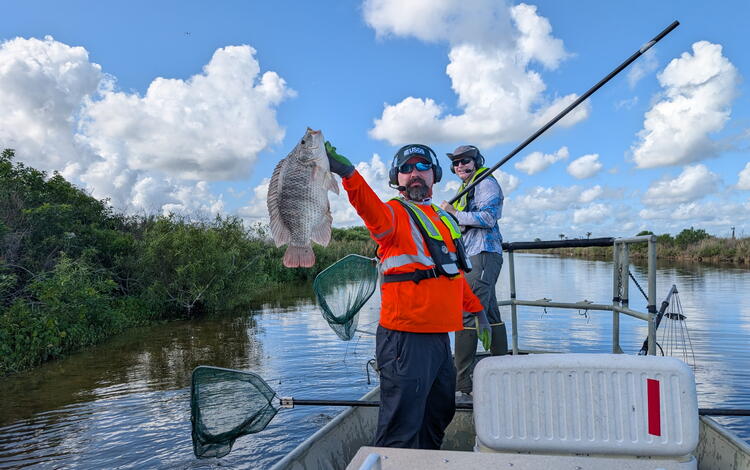 two people in life vests stand in a boat - one holds a fish while the other holds a dipnet