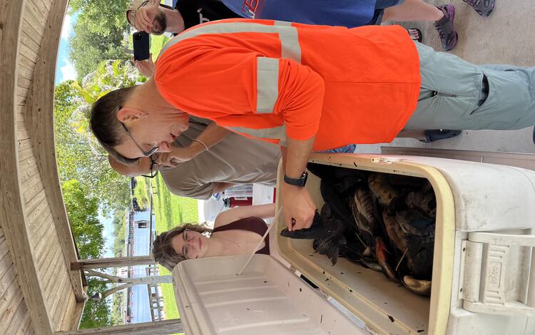 A group of people stand around a cooler filled with nonnative fish as a man in an orange shirt gives identification guidance
