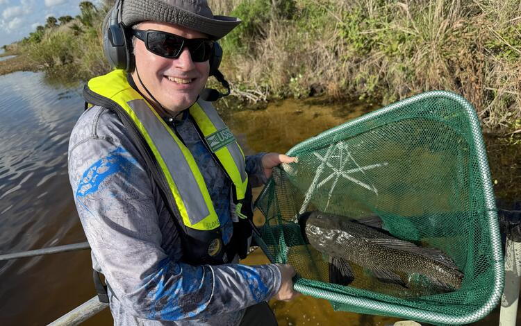a man holding a fish in a net stands with water and vegetation seen in the background