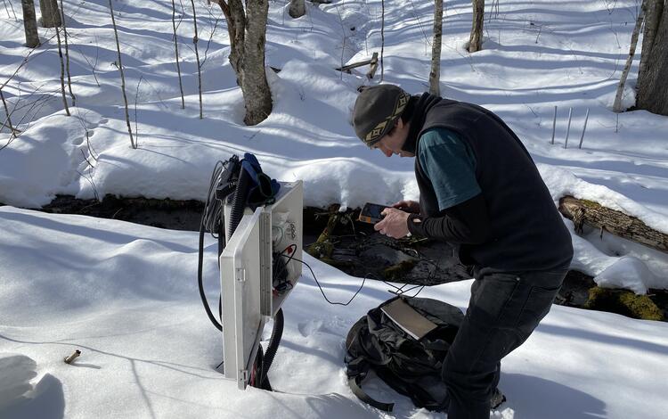 A man in the woods in the snow looks at a data logger.