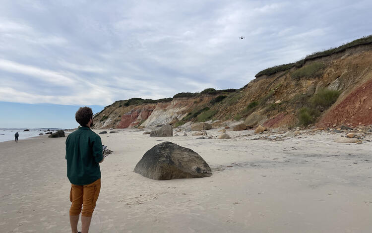 person standing on sandy beach with control in hand looking at drone in the sky over cliffs