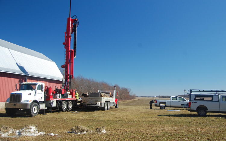 Several vehicles and a large underground drilling rig with a few people in a field beside a building. 