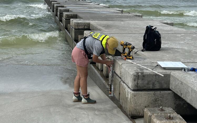 A scientist installs a piece of equipment along the base of a long, concrete pier leading out the ocean. 