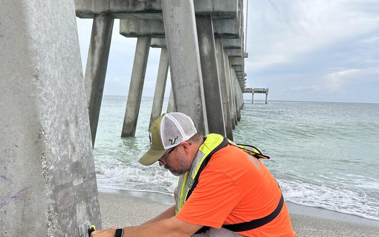 A scientist installs a piece of equipment along the base of a long pier leading out the ocean. 