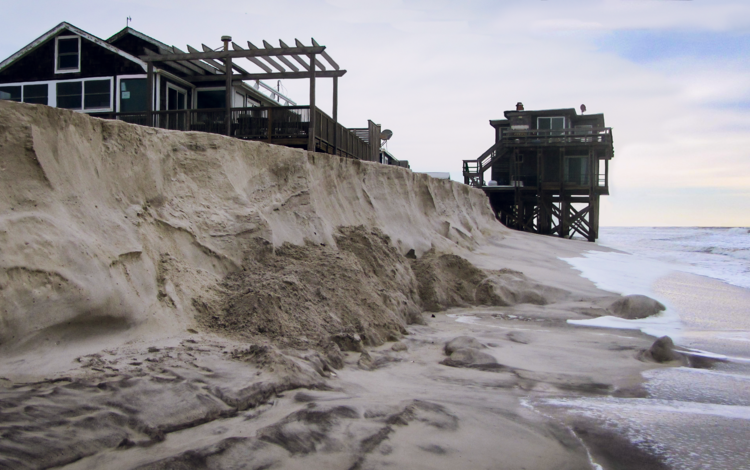 a sandy coastline with heavily eroded dunes with two houses on stilts nearby
