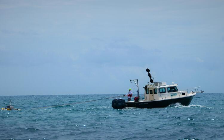 a green and white research boat on the water towing an object with two yellow pontoons