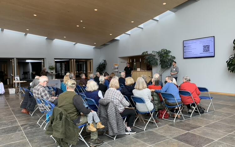 Photo of a seated group of people in a room, facing a screen. Presenters are at the front of room, near the screen.