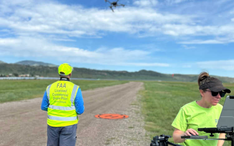 two scientists in bright yellow shirts stand on an airplane landing strip in a field, with a small UAS in the air above them
