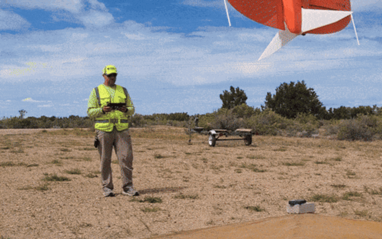 A man in neon yellow gear holds a remote control, an orange drone rises up and lands on a helipad in front of the man