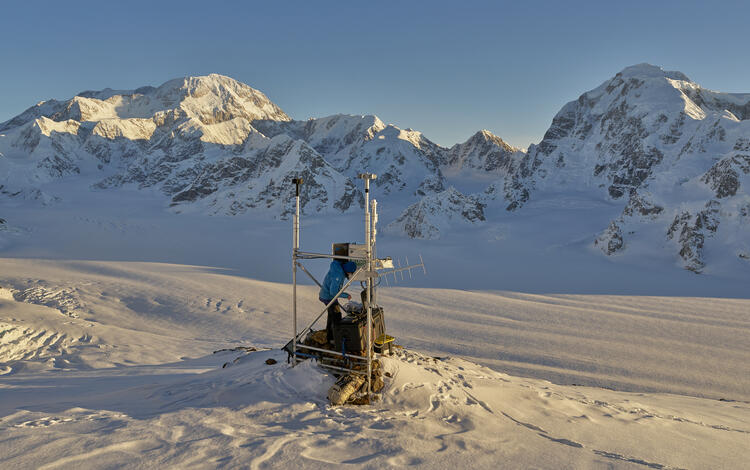 Person in a blue jacket working at a weather station in the snow with mountains in the background 