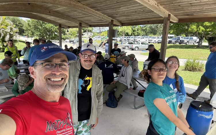 Four people stand in the foreground taking a selfie-style photo, with many people sitting on metal park benches under a large pavilion in the mid-ground, and a road and parking lot in the background
