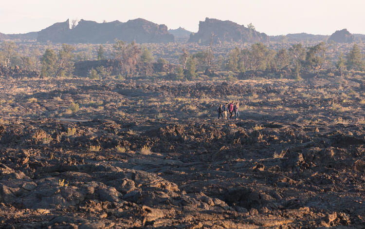 four people walk across a lava bed, trees and rocky hills in the background