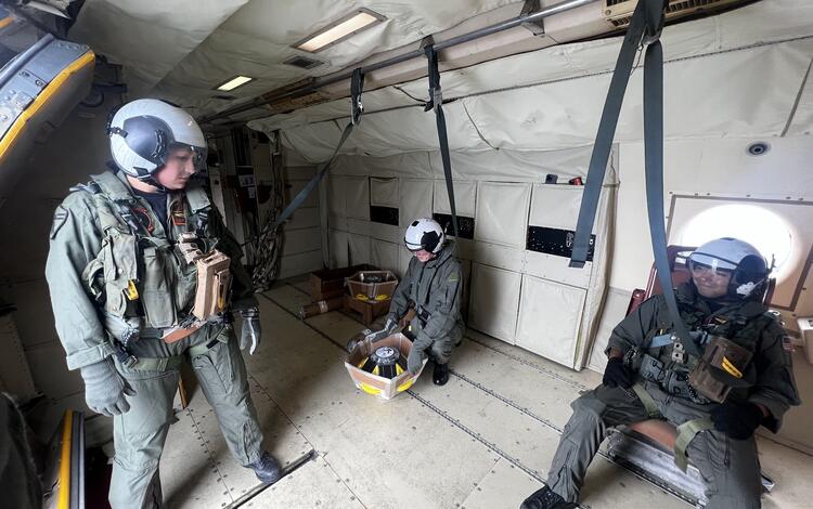 Three people inside a plane wearing flight suites and helmets, two are seated, one is standing looking at them