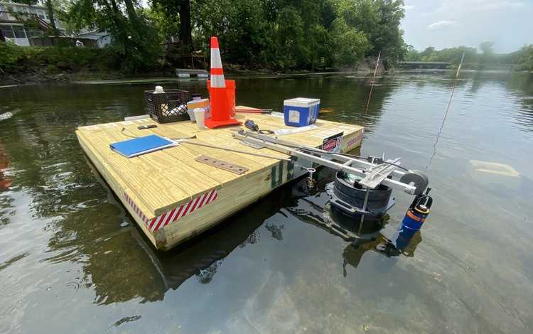 Orange traffic cone, sample storage box, and a notebook sit on a wooden platform in the river with the Bacteria sampler.