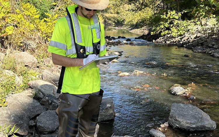 Scientist records conditions of river while standing next to it with a highway overpass in background