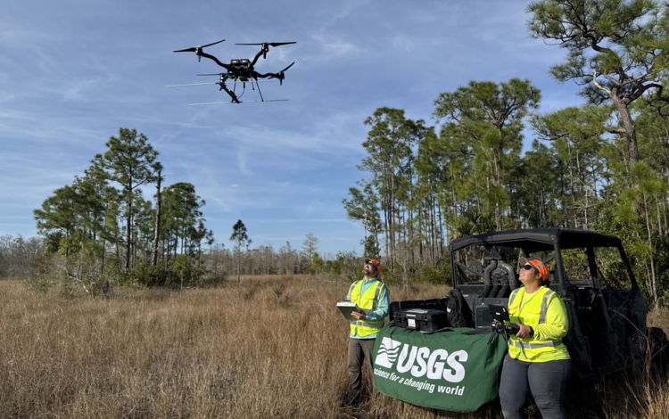 two people in yellow vests lean against an OHV, look up at drone flying above them, grasses and trees in background