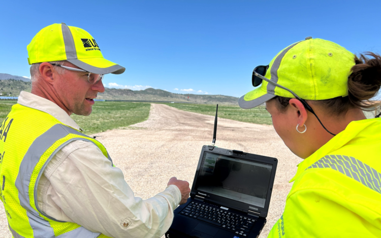 two people in bright yellow USGS gear chat outside while looking at a small computer screen