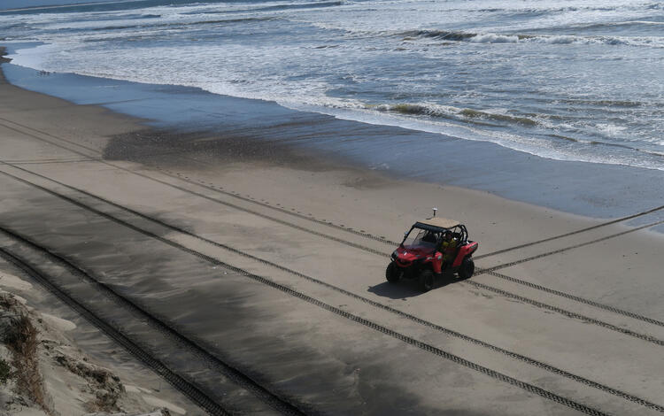 View down to beach from coastal bluff looking at an all-terrain vehicle that has left very precise, perpendicular tire tracks.