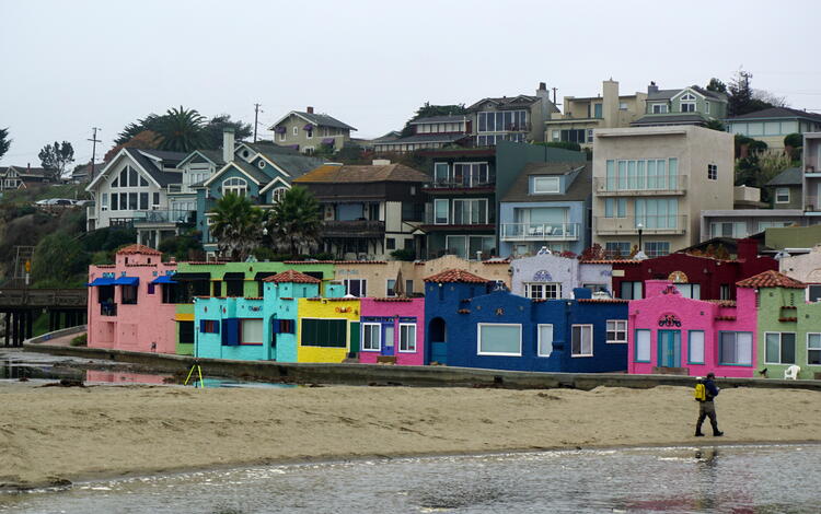 Man walks along the sandy edge of a river with very colorfully painted apartments in background.