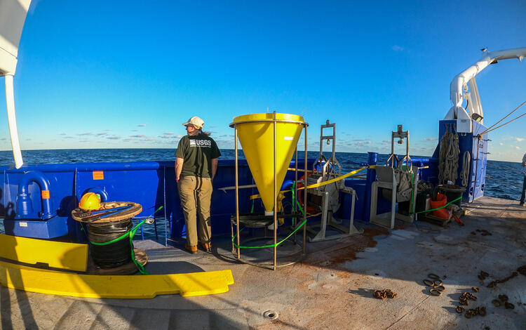 Woman stands besides scientific equipment on the deck of a ship.