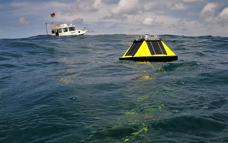 A yellow buoy with solar panels floats in the water with a boat in the background