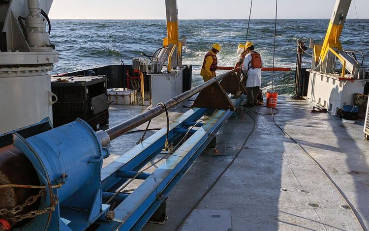 On the stern of a ship, a crew works on a large piece of equipment that lies on the deck in a frame to hold it securely.