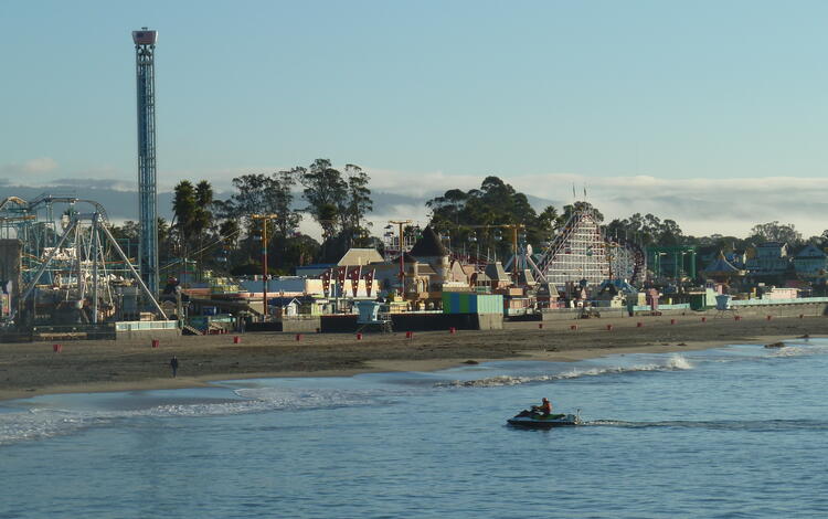 In foreground, man rides a personal watercraft in the water near a beach, in background is an amusement park with carnival rides