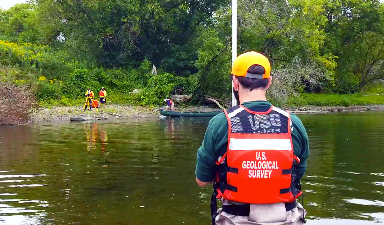 USGS employee stands in the river with a prism while a point measurement is taken with a station located on the river bank.